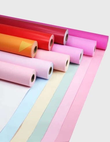 Rollo de celofán liso de alta transparencia y acabado brillante, rodeado de diversas presentaciones en pliegos, bolsas y rollos de colores vibrantes, destacando su textura suave sobre fondo blanco.